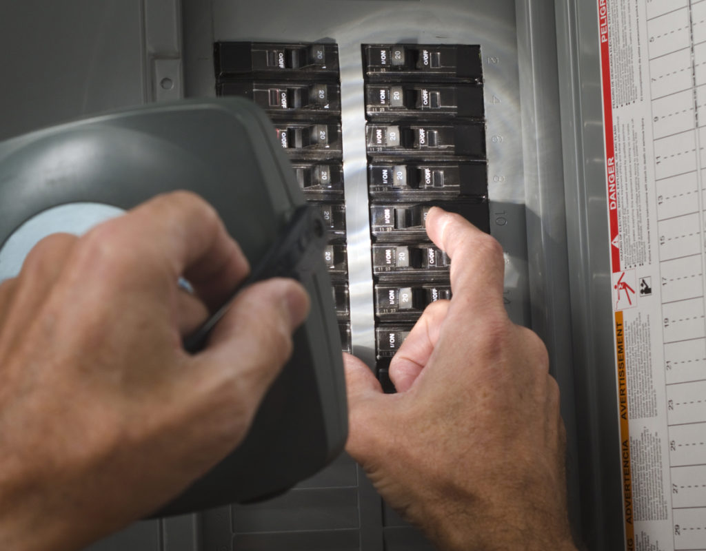 Image of a man's hands and a circuit breaker panel. He is holding a flashlight in one hand, flipping a switch with the other.